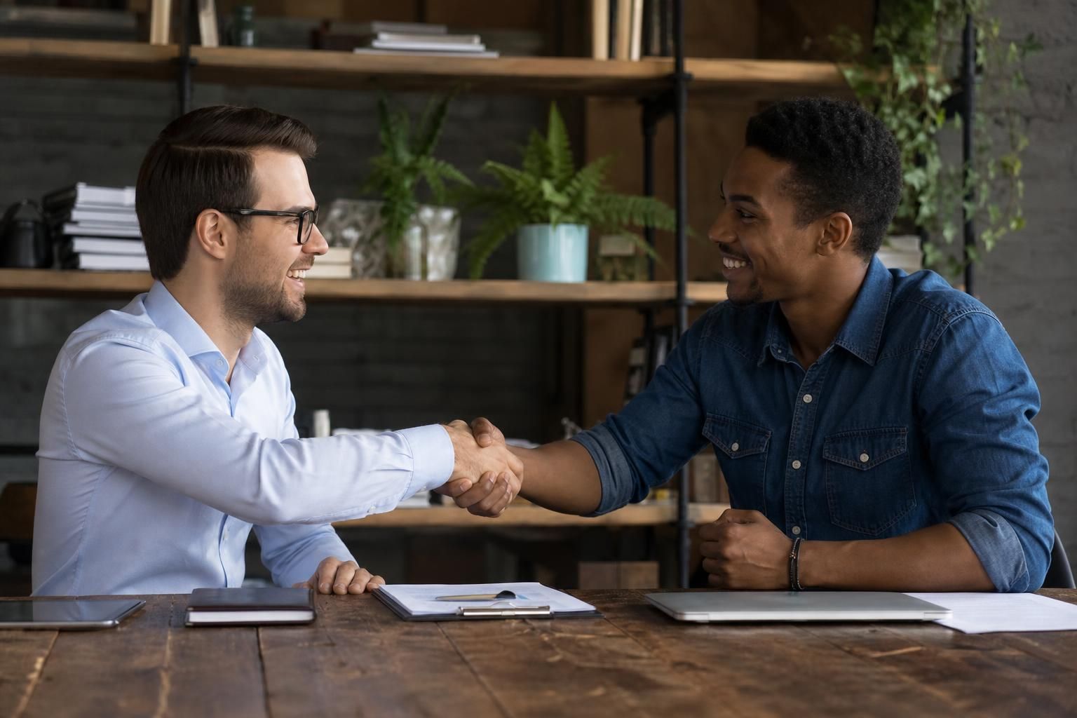 Two men shake hands across a desk with a laptop and clipboard